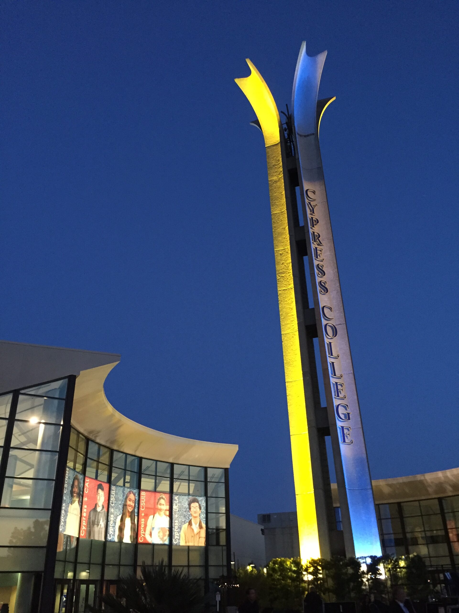 Cypress College Campanile, a spire, lit up in blue and gold. A building is to the left.