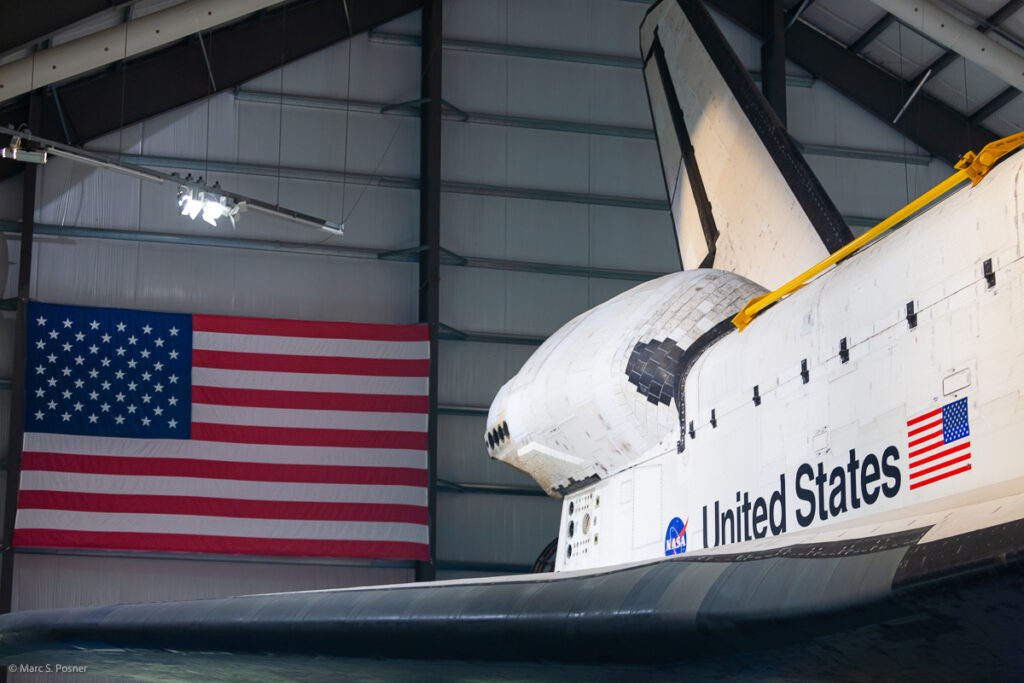 Photograph of space shuttle Endeavour showing the the NASA meatball logo, the words United States, and the US flag from the right side of the orbiter. A US flag also hangs in the background.