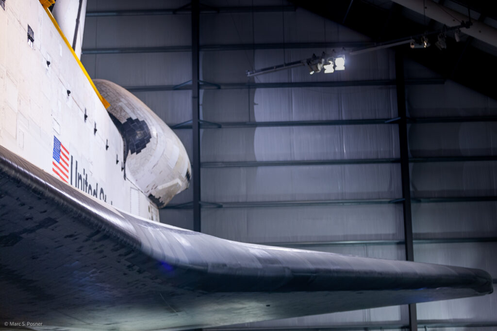 Photograph of space shuttle Endeavour showing part of the left side of the orbiter. Visible is the leading edge of the left wing, the US flag, the left OMS pod and parts of the rudder and cargo bay doors.