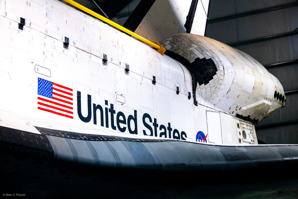 Photograph of space shuttle Endeavour showing part of the left side of the orbiter. Visible is the leading edge of the left wing, the US flag, the words United States, the left OMS pod and parts of the rudder and cargo bay doors.