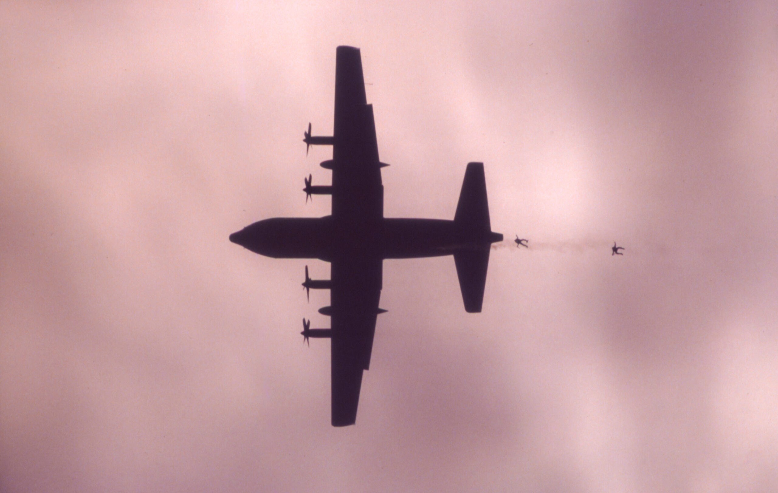 A C-130 aircraft as seen looking straight up. It is silhouetted against a grayish sky. Two paratroopers can be seen jumping from the plane and trailing behind the tail.