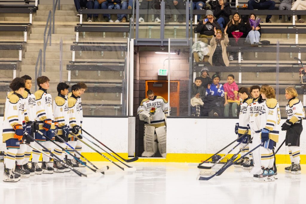 an ice hockey player waits to enter the rink in a doorway as he holds flowers and teammates form a line for him to skate past