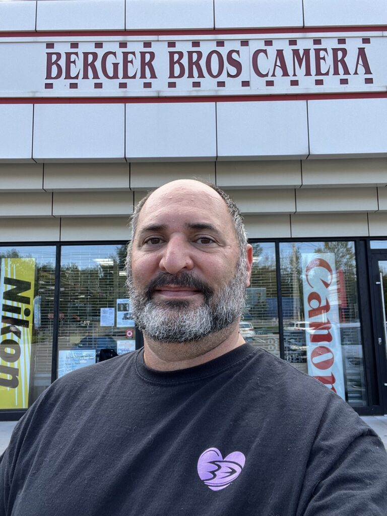 A man with a partially grey bears stands in front of a store with a sign reading Berger Bros Camera and signs in the window reading Canon and Nikon