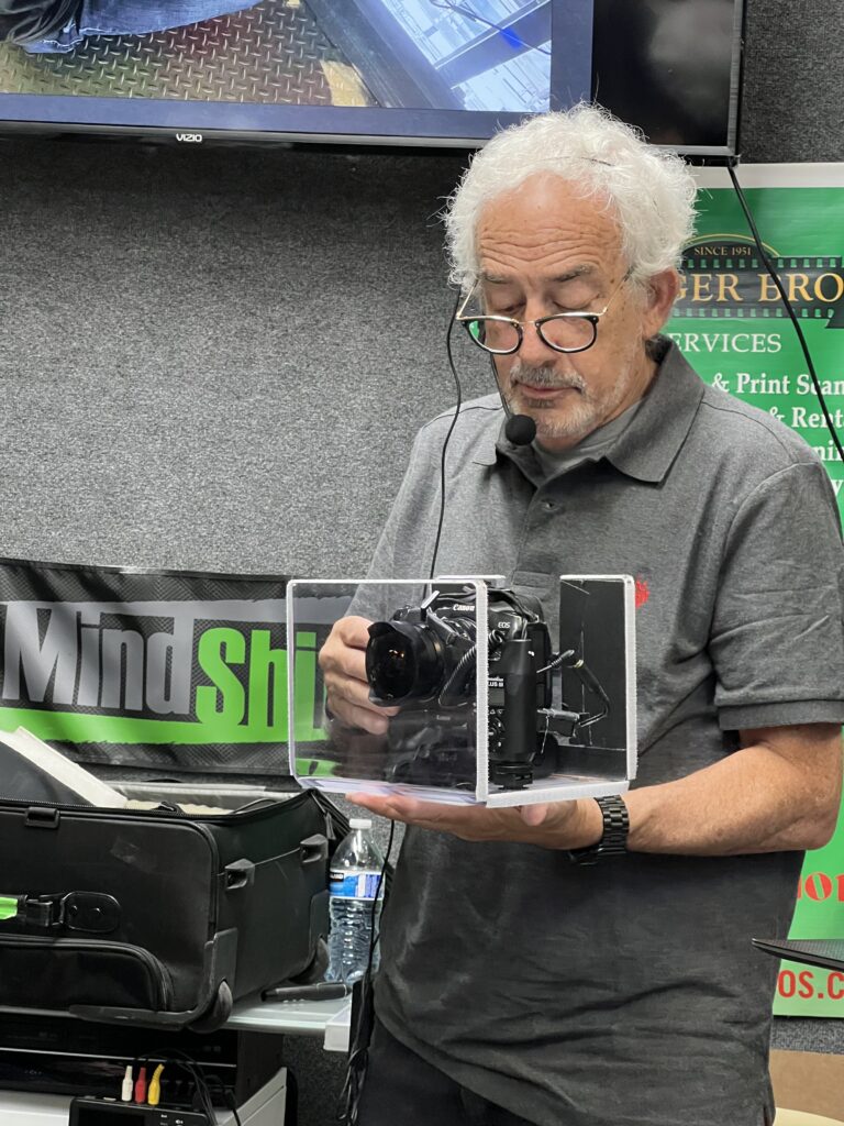 A man with grey hair holds up a camera mounted inside a transparent acrylic box.