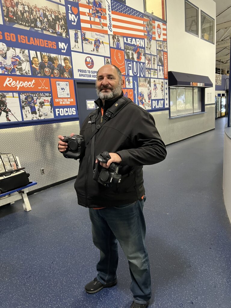 A man with a grey beard stands inside an ice arena holding two professional cameras